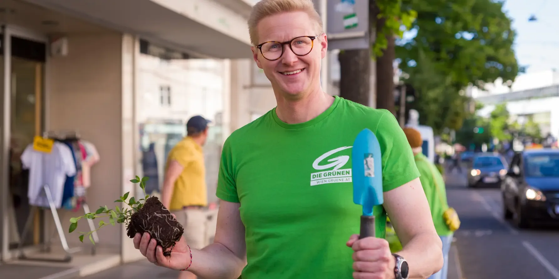 Pascal Riepl hält eine Schaufel in der linken Hand und eine Pflanze in der rechten. Er guckt in die Kamera und lächelt.