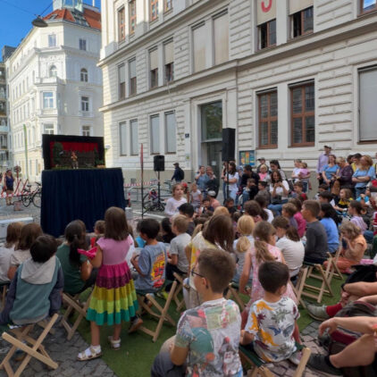 Eine Gruppe von Kindern sitzen auf der Straße und gucken den Kasperl.